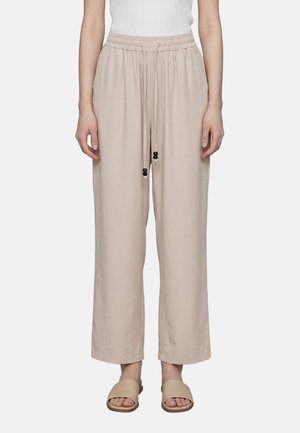 Beige locker sitzende Hose mit elastischem Bund und Kordelzug, getragen mit beigen offenen flachen Sandalen an einer stehenden Person.