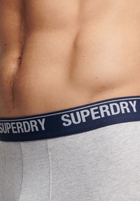 Grijze boxershorts met een marineblauwe elastische tailleband waarop de tekst "SUPERDRY" in het wit is gedrukt, met nadruk op textuur en bandontwerp.
