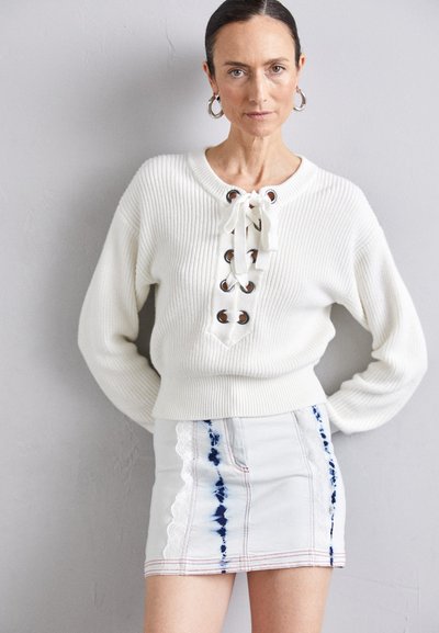 Pull en maille blanche à côtes, avec un détail à lacets et des œillets en métal. Associé à une jupe en denim claire ornée de motifs tie-dye et d'accents en dentelle.