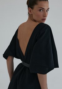 Czarna sukienka z głębokim dekoltem na plecach i szerokimi rękawami; przewiązana w talii. Materiał wydaje się teksturowany, z miękką i zwiewną sylwetką.