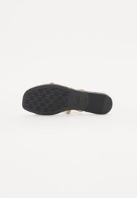 Flat sandal with a textured black rubber sole, featuring a metallic gold strap with decorative elements on the upper.