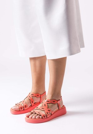 Pieds portant des sandales rose corail avec des brides festonnées et des boucles argentées, associés à un pantalon blanc à jambes larges coupé au-dessus des chevilles.