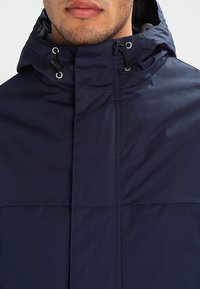 Giacca con cappuccio blu navy realizzata in un tessuto liscio e strutturato, dotata di cordini regolabili, chiusure a scatto e zip frontale.