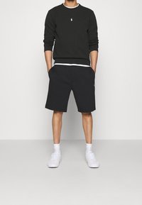 Svart sweatshirt med vit krage och liten logotyp, matchad med mörka shorts och vita sneakers. Modellen står med händerna i fickorna.