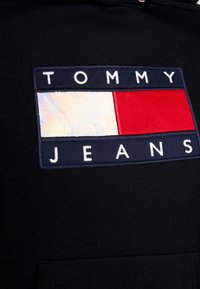 Sudadera de algodón negra con un gran parche con "TOMMY JEANS" en bordado blanco, acentos rectangulares en rojo y blanco sobre un fondo azul marino.