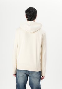 Crème-kleurige hoodie met een zachte textuur, voorzien van een grote capuchon en geribbelde manchetten. Draagt met blauwe jeans, gezien vanuit de achterkant.