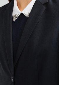 Svart blazer med texturerad tyg, slagkrage och vit krage med kristalldekoration längs kanten.