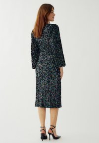 Juodas pailgas sequinais užtiestas suknelė ilgomis rankovėmis, iki kelių ilgio, su tekstūruota paviršiumi. Dėvima su juodomis aukštakulnėmis bateliais su raišteliais.