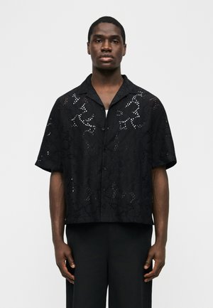 Moški, ki nosi ohlapno črno srajco s kratkimi rokavi z gumbi in cvetličnim izrezanim vzorcem ter črne hlače, stoji pred enobarvnim belim ozadjem.