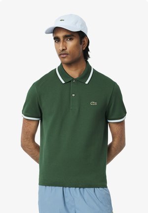 Grön polo t-shirt med klassisk krage, vit kant på ärmarna och en logotyp på bröstet. Bärs med en ljusblå keps. Slät tygtextur.