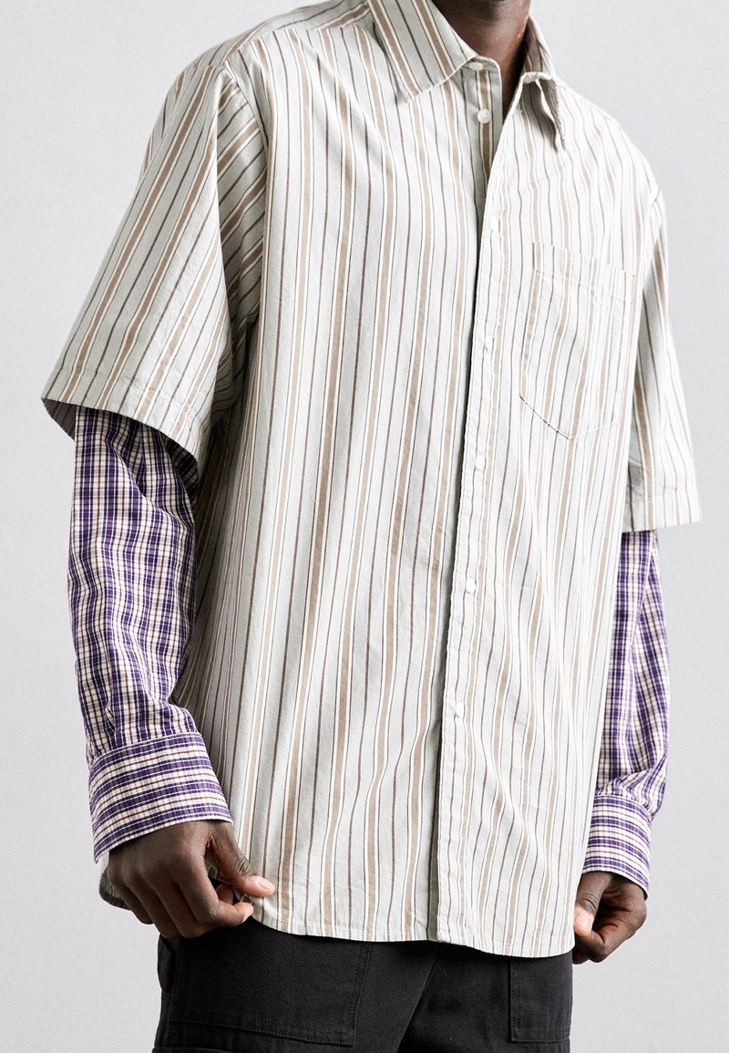 Mężczyzna w warstwowych koszulach: długie rękawy w fioletową kratę pod krótkim rękawem beżowej koszuli w pionowe paski, w połączeniu z czarnymi spodniami.