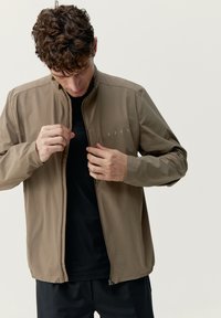 Ljusbrun, lättviktsjacka med hög krage, frontdragkedja och subtilt varumärkeslogotyp. Bärs över en svart t-shirt med lång ärm.