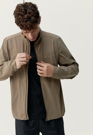 Veste léger couleur marron clair, avec un col haut, une fermeture éclair à l'avant et un logo de marque discret. Portée par-dessus un t-shirt noir à manches longues.