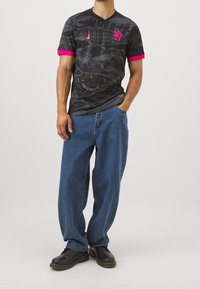 Maglietta nera a scollo a V con dettagli rosa, caratterizzata da un motivo testurizzato e un logo. Abbinata a jeans blu larghi e scarpe nere.