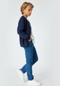 Sudadera azul marino con cierre, camiseta blanca, jeans azules y zapatillas blancas con detalles grises. Presenta un corte relajado y puños acanalados.