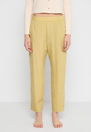 Osoba ubrana w jasnożółte proste spodnie oraz beżowy krótki, zapinany na guziki top z długim rękawem, stojąca boso na jednolitym tle.