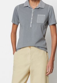 Grå kortärmad polo med kängurukrage, femknappslå, och bröstficka, i kombination med ljusbeige byxor.