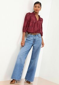 Femme portant un blouse bordeaux texturé, un jean large bleu clair avec une ceinture marron et des escarpins pointus marron, se tenant contre un mur blanc.