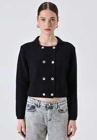 Cardigan nero corto in maglia testurizzata con colletto, dotato di grandi bottoni argentati sul davanti. Abbinato a jeans grigi chiari e strappati.