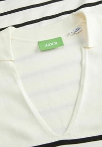 Suéter de rayas crema con escote en V, rayas horizontales negras y una etiqueta verde que dice "JJXX" en el cuello. Textura suave.