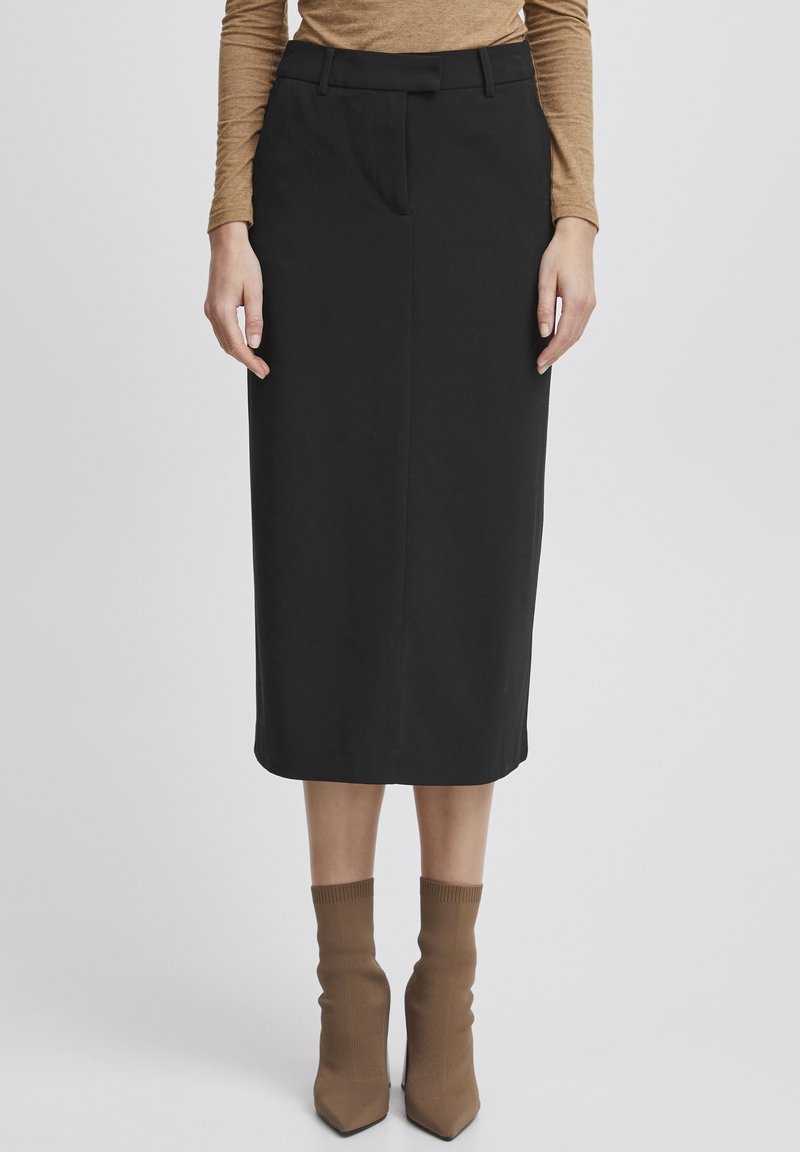 Jupe crayon noire à texture lisse et longueur mi-mollet, dotée d'une taille cintrée et de plis avant subtils. Portée avec des bottines marron clair.