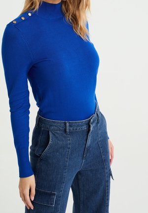 Vrouw draagt een helderblauwe trui met lange mouwen, schouderknopen en een hoge taille donkere spijkerbroek met voorzakken.