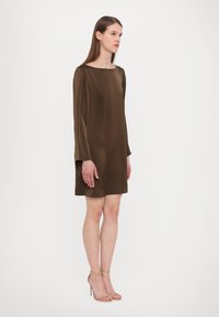 Ruskea silkkimekko, jossa on venepääntie, pitkät hihat ja A-linjainen siluetti, yhdistettynä beigeihin remmisandaaleihin. Suora pinta, minimalistinen muotoilu.