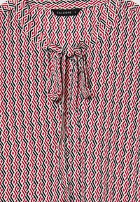 Blusa a fantasia color crema, con un motivo geometrico a diamante rosso e nero, dotata di un dettaglio a fiocco sul colletto e una texture del tessuto liscia.