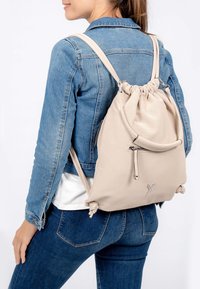 Mochila de tela perforada beige claro con correas ajustables, cierre de cuerda y bolsillo con cremallera en la parte frontal, llevada sobre una chaqueta de mezclilla.