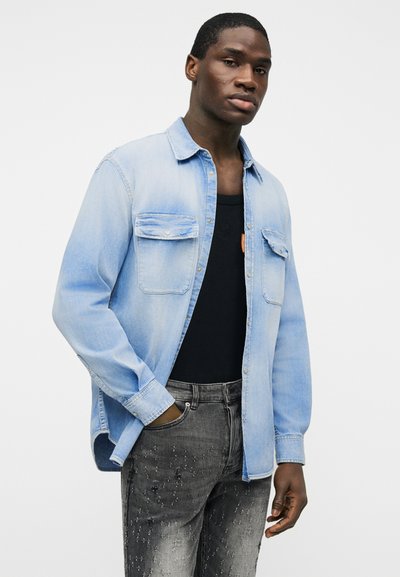 Νέος άνδρας που φοράει ανοιχτό μπλε τζιν πουκάμισο πάνω από μαύρο αμάνικο και φθαρμένο γκρι τζιν, στέκεται με ένα χέρι στην τσέπη.