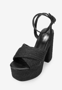 Sandalia de plataforma negra que presenta un upper de tela trenzada, diseño de tiras cruzadas y tacón cuadrado grueso. Incluye tira ajustable en el tobillo.