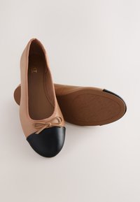 Ballerines beiges avec bouts noirs et petits nœuds sur le devant, une chaussure debout et l’autre montrant la semelle avec des motifs antidérapants.