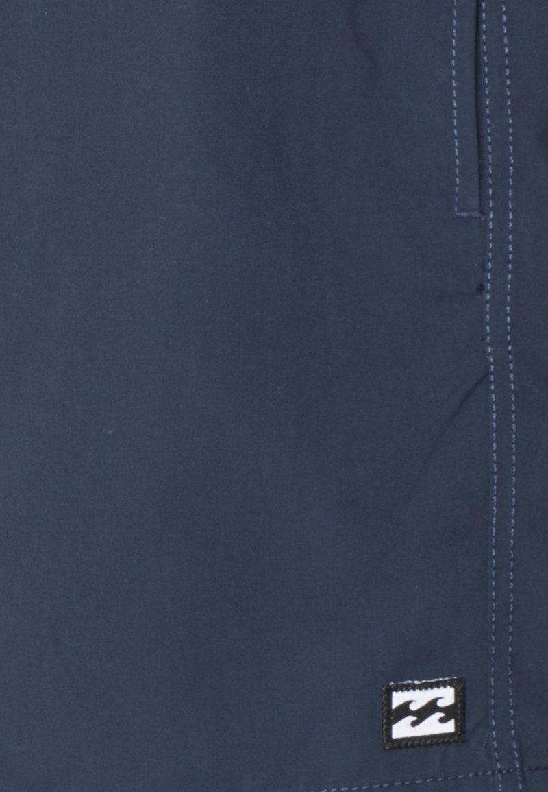 Navyblauwe stof met een gladde textuur, voorzien van contrasterende witte stiksels en een zwart label met een golflogo in de rechterbenedenhoek.