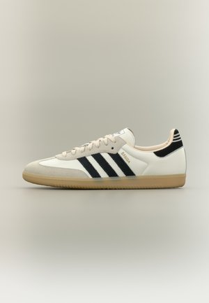 Witte en beige Adidas Samba lage sneaker met zwarte strepen en een gumkleurige zool, zijaanzicht op een neutrale achtergrond.
