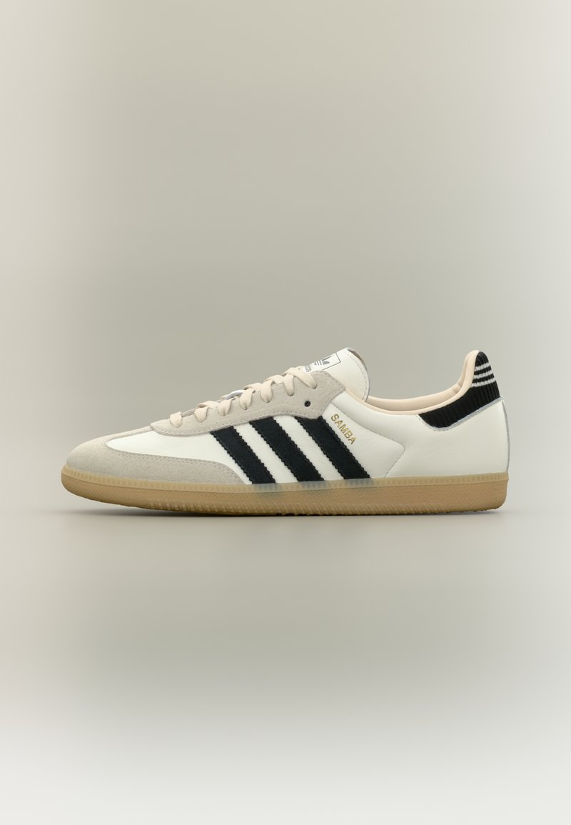 Άσπρο και μπεζ χαμηλό παπούτσι Adidas Samba με μαύρες ρίγες και σόλα από καουτσούκ, εμφανιζόμενο σε πλευρική όψη σε ουδέτερο φόντο.