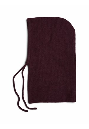 Mujer joven con una capucha de punto de color púrpura oscuro con cordones y un abrigo negro con botones sobre un fondo claro y liso.
