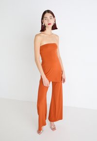 Pánt nélküli, testhezálló top terrakotta színben, széles szárú nadrággal párosítva. Sima, rugalmas anyag, elegáns kivitel. Ezüst színű magassarkú szandál.