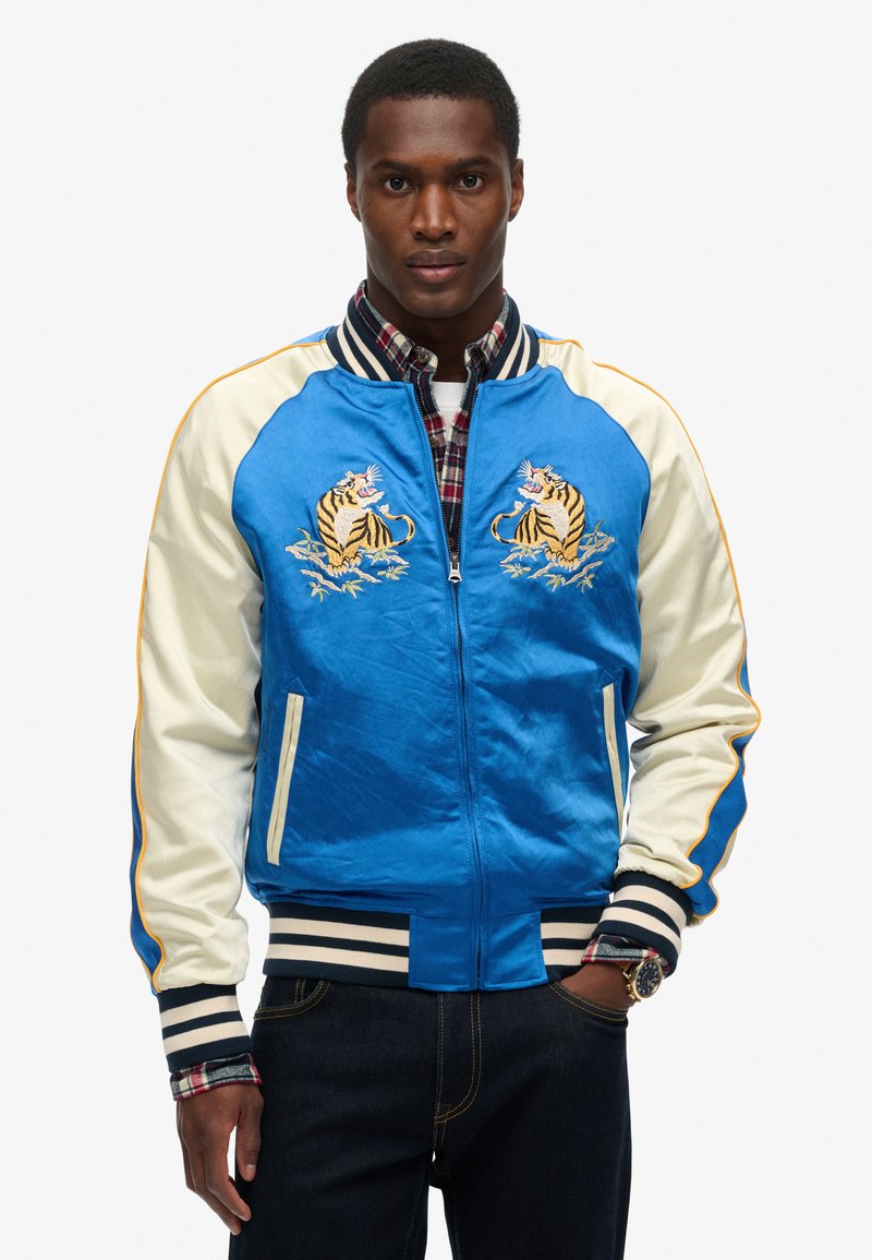 Homme portant une veste bomber satin bleu et crème avec des tigres brodés, une chemise à carreaux en dessous, un jean foncé et une montre, se tenant contre un fond blanc.