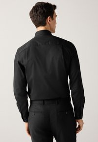 Camicia nera a maniche lunghe con schiena strutturata, polsini con bottoni e vestibilità sartoriale. Il tessuto appare liscio con una minima trama.