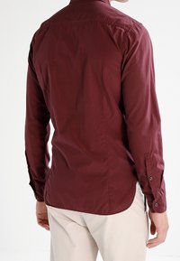 Muž oblečený v tmavě červené košili s dlouhým rukávem a světlých béžových kalhotách, zobrazený zezadu na bílém pozadí.