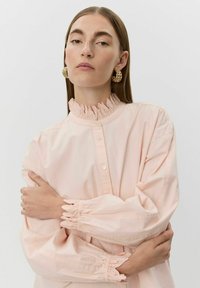 Svetloružová blúzka s vysokým volánovým golierom a nariasenými rukávmi. Má zapínanie na gombíky vpredu a mierne nafúknuté rukávy pre pohodlný strih.