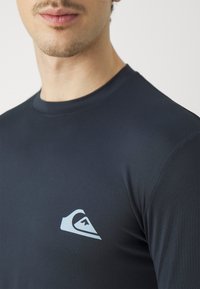 Námornícky modrý priliehavý rash guard vyrobený z hladkého materiálu, s svetlomodrým logom na ľavej strane hrude a s dizajnom okrúhleho výstrihu.