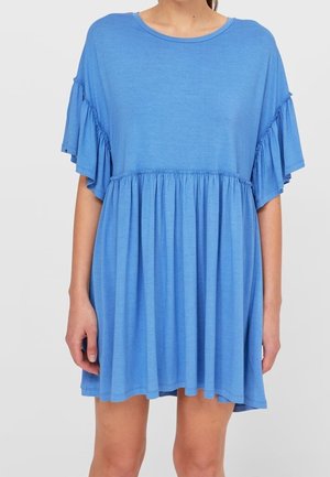 Vrouw draagt een losse, korte blauwe jurk met geplooide, gelaagde mouwen en een geplooid tailledetail, staand tegen een effen achtergrond.