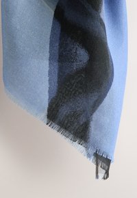 Woven scarf in blue and black with a gradient design. Soft texture, fringed edges, and abstract patterns visible.