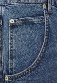 Close-up of a blue denim jeans pocket with orange stitching and metal rivets on textured fabric.