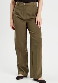 Vrouw draagt olijfgroene rechte broeken en zwarte ballerina's, staand tegen een effen witte achtergrond.