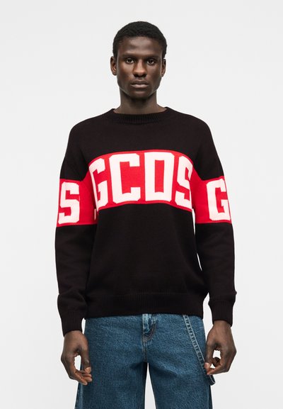 Άνδρας που φοράει μαύρο πουλόβερ με έντονα κόκκινα και λευκά γράμματα "GCDS" στο στήθος και στα μανίκια, συνδυασμένο με μπλε τζιν.