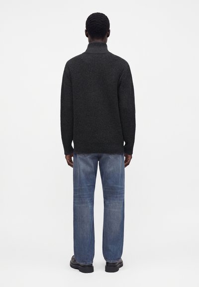 Personne debout de dos portant un pull à col roulé gris foncé, un jean bleu et des chaussures noires sur un fond blanc uni.