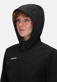Zwarte softshell jas met een capuchon, voorzien van een ritssluiting aan de voorkant, een gestructureerde stof en de merknaam "MAMMUT" in het wit geborduurd op de borst.