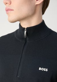 Sudadera negra con cremallera y cuello alto, diseño de punto texturizado y un logo blanco "BOSS" en el pecho. Detalle de cremallera plateada visible.